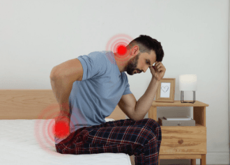 man sitting on edge of bed holding head and back due to pain